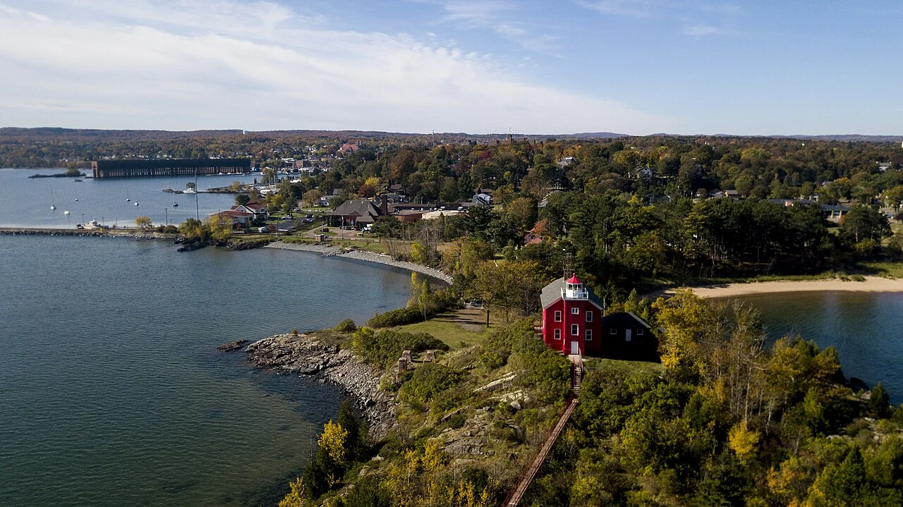 Aerial view of the red Marquette Harbor Light on a wooded point, with Marquette in the background.